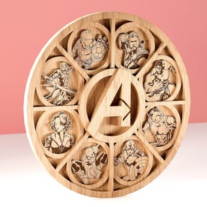 Puede incluir: Una obra de arte mural circular de madera con retratos cortados con láser de personajes de los Vengadores. El diseño incluye a Iron Man, Thor, Hulk, Capitán América, Viuda Negra y Spider-Man. El elemento central es la letra "A".