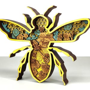 Peut inclure: Sculpture en bois en forme d'abeille à plusieurs couches, avec des accents jaunes, marron et bleus. L'abeille présente des découpes complexes de fleurs, de nids d'abeilles et le mot "Honey". Les ailes et le corps sont détaillés.