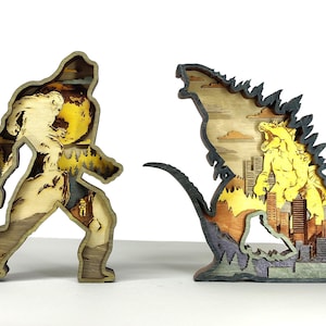 Op de afbeelding: Twee houten gelaagde sculpturen: een van Bigfoot en de andere van Godzilla. De Bigfoot-sculptuur is in tinten van bruin, geel en beige. De Godzilla-sculptuur is in tinten van geel, bruin en grijs, met een stadsgezicht op de achtergrond.