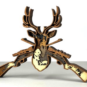 Peut inclure: Une œuvre d'art en bois multicouche représentant une tête de cerf avec des bois, deux fusils et une scène de forêt. L'œuvre est principalement marron avec des accents de bois plus clairs, créant un effet 3D. Idéal pour la décoration rustique ou sur le thème de la chasse.