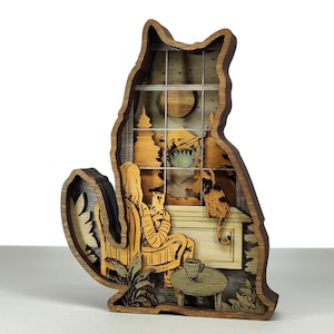 以下が含まれることがあります： 多層木製の猫型装飾品。内部には、人、猫、窓、カップのテーブルがある居心地の良いシーンが描かれています。この作品は、さまざまな茶色とタン色の木材で作られており、暖かく居心地の良い美しさを生み出しています。