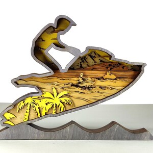 Pu&ograve; includere: Scultura in legno a strati raffigurante una persona che fa jet ski. La scultura presenta palme gialle, un cielo giallo e una base grigia. La moto d'acqua &egrave; grigia con interni gialli e l'acqua &egrave; un mix di giallo e marrone.