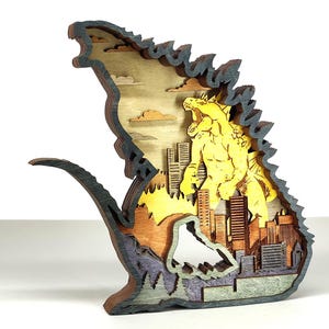 Op de afbeelding: Een meerlaagse houten sculptuur die een gele monsterfiguur boven een skyline afbeeldt. De sculptuur heeft verschillende tinten bruin, grijs en geel, met een gedetailleerde stadsgezicht en een gestileerd monsterontwerp.