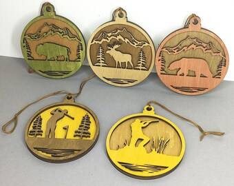 Pode incluir: Uma coleção de cinco enfeites de madeira com designs temáticos da natureza. Cada enfeite é circular com um design recortado apresentando um bisonte, um alce, um urso, um golfista e um caçador. Os enfeites têm uma cor de madeira natural com detalhes em verde, amarelo e laranja, e uma corda para pendurar.