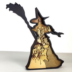 May include: A layered wooden witch figure with a broom. The witch is black and the interior is a light brown. The figure has a witch hat and is holding a broom. The interior shows a scene with people. The figure is standing on a white surface.