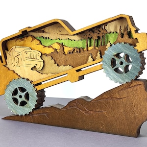 Op de afbeelding: Een gelaagde houten sculptuur die een jeep voorstelt die een berg beklimt. Het kunstwerk toont een gedetailleerd landschap met bomen, bergen en een jeep-ontwerp. De wielen zijn lichtblauw en het kleurenschema is in natuurlijke houttinten.