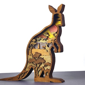 Puede incluir: Escultura de madera de un canguro con un diseño en capas. El canguro es marrón con una escena interior detallada que presenta árboles, una puesta de sol y otros animales. La escultura mide aproximadamente 25 cm de altura.