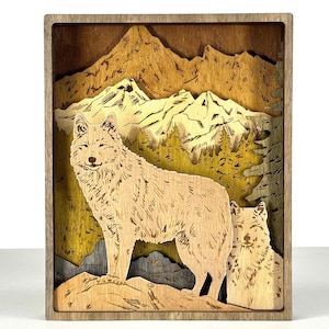 May include: A wooden layered art piece featuring a wolf and a pup in a mountain landscape. The artwork uses natural wood tones to depict the animals, mountains, and trees. The frame is a darker wood, creating a shadow box effect.