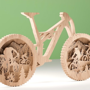 May include: A wooden mountain bike sculpture with intricate cutouts. The wheels feature scenes of cyclists riding through a mountain landscape with trees and peaks. The bike is made of light-colored wood and stands against a green background.