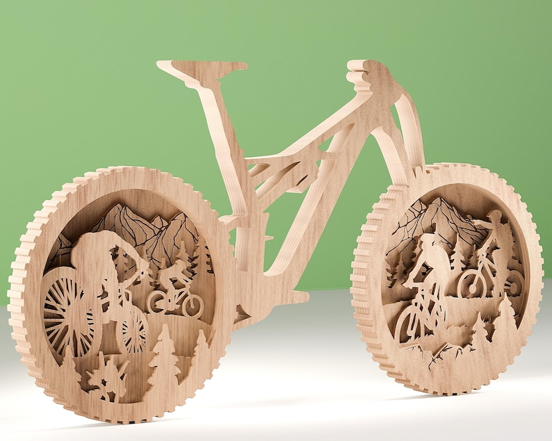 May include: A wooden mountain bike sculpture with intricate cutouts. The wheels feature scenes of cyclists riding through a mountain landscape with trees and peaks. The bike is made of light-colored wood and stands against a green background.