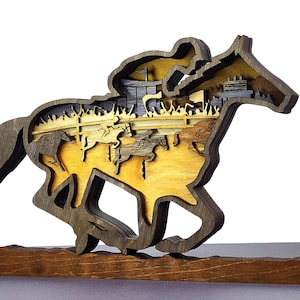 May include: A multi-layered wooden sculpture depicting a horse race. The sculpture features a horse and jockey in motion, with other horses and riders visible in the background. The piece is made of various shades of brown and gold wood, mounted on a rectangular base.