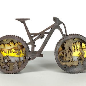 May include: A wooden mountain bike sculpture with intricate cutouts depicting cyclists in a landscape. The wheels feature detailed scenes of riders, trees, and mountains, with a yellow and brown color scheme. The bike is a dark brown color.