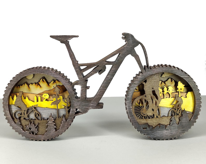 May include: A wooden mountain bike sculpture with intricate cutouts depicting cyclists in a landscape. The wheels feature detailed scenes of riders, trees, and mountains, with a yellow and brown color scheme. The bike is a dark brown color.