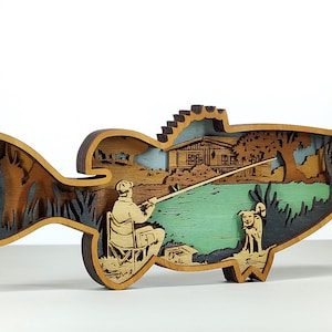 May include: A layered wooden art piece in the shape of a fish. The artwork depicts a fisherman seated in a chair, fishing near a lake, with a dog. A cabin and trees are in the background. The piece is made of various shades of brown, green, and blue.