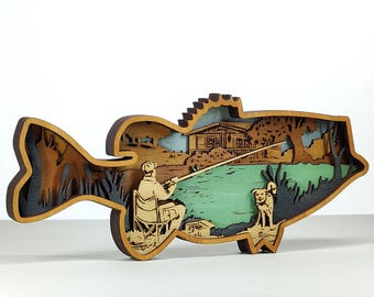 May include: A layered wooden art piece shaped like a fish. It shows a fisherman fishing by a lake, with a dog, a cabin, and trees. The artwork uses shades of brown, green, and blue. The piece is a decorative item.