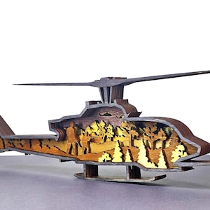 Może przedstawiać: Drewniany model helikoptera o warstwowej konstrukcji. Wnętrze przedstawia krajobraz z drzewami i postaciami. Zewnętrzna część jest ciemnobrązowa, a wewnętrzne warstwy mają jaśniejsze odcienie drewna. Model posiada wirnik i ogon.