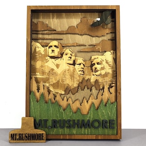 May include: Wooden layered art piece depicting Mount Rushmore with an eagle in flight. The artwork features a brown wooden frame, with the mountain faces carved in detail. The base has green and brown trees and grass. The text "MT. RUSHMORE" is at the bottom.