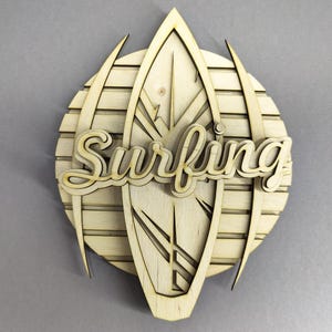 May include: Wooden wall art featuring a surfboard design with the word "Surfing" in a cursive font. The artwork is made of light-colored wood and has a layered design, with a circular background and pointed accents.