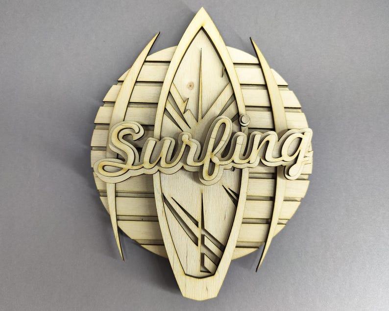 May include: Wooden wall art featuring a surfboard design with the word "Surfing" in a cursive font. The artwork is made of light-colored wood and has a layered design, with a circular background and pointed accents.