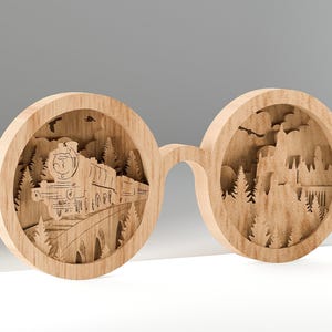 Puede incluir: Objeto de arte de madera con forma de gafas. Una lente muestra una escena de tren, la otra un castillo. La madera clara tiene intrincados detalles cortados con láser de árboles, un tren y un castillo. Un objeto decorativo.