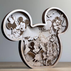 May include: A wooden, layered art piece in the shape of Mickey Mouse's head. The artwork features engraved images of Mickey, Minnie, and other Disney characters. The word "Mickey" is written in script. The piece is made of light-colored wood.