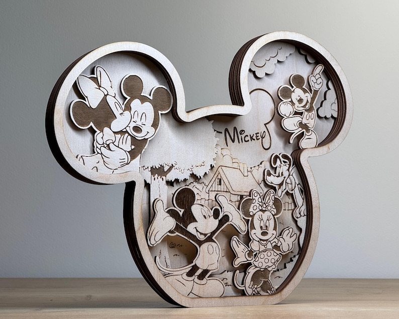 May include: A wooden, layered art piece in the shape of Mickey Mouse's head. The artwork features engraved images of Mickey, Minnie, and other Disney characters. The word "Mickey" is written in script. The piece is made of light-colored wood.
