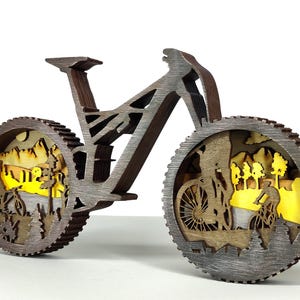 May include: A wooden bicycle sculpture with intricate cutouts depicting cyclists and a mountain landscape. The wheels feature detailed scenes with yellow backlighting, creating a layered effect. The frame is a dark brown color.