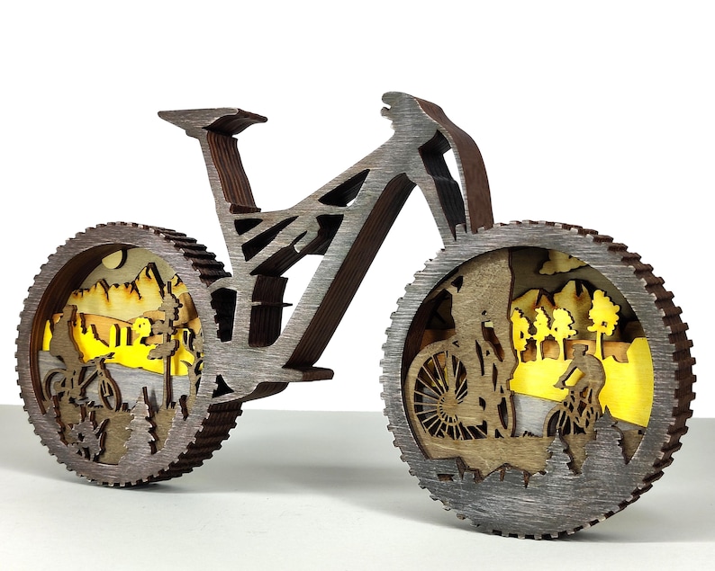 May include: A wooden bicycle sculpture with intricate cutouts depicting cyclists and a mountain landscape. The wheels feature detailed scenes with yellow backlighting, creating a layered effect. The frame is a dark brown color.