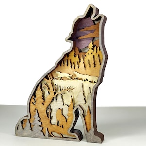 May include: A layered wooden sculpture of a howling wolf, featuring a forest scene with trees, a moon, and other wolves. The sculpture is made of various shades of brown and gray wood, creating a 3D effect. The wolf is in a sitting position.