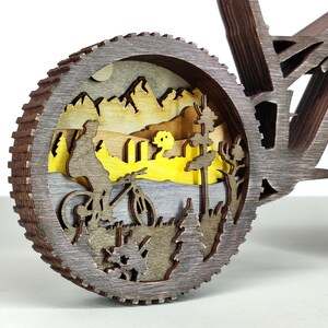 May include: A wooden bicycle sculpture with a detailed wheel design. The wheel features a layered landscape scene with a cyclist, mountains, and trees. The sculpture is made of various shades of brown and gray wood.