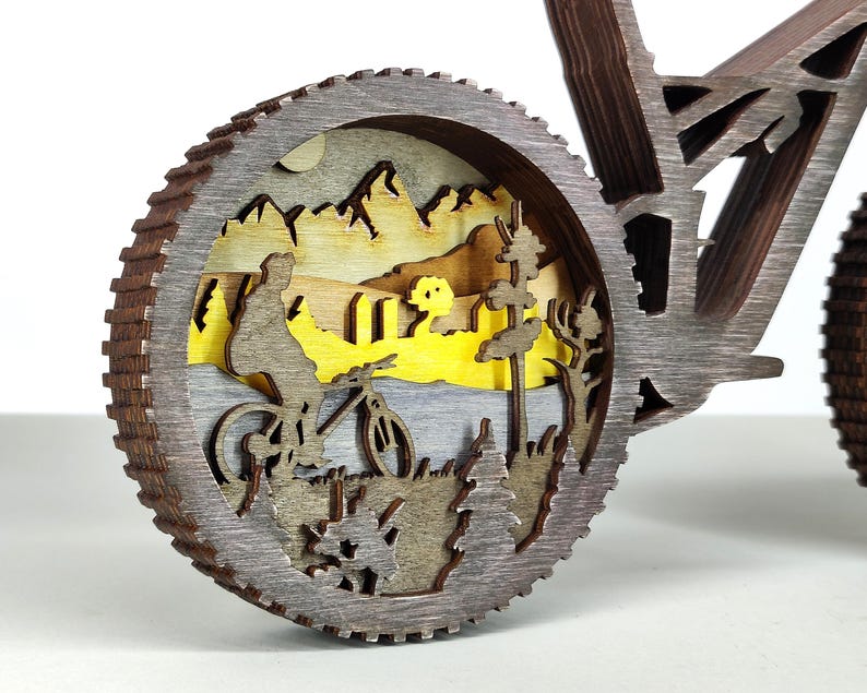 May include: A wooden bicycle sculpture with a detailed wheel design. The wheel features a layered landscape scene with a cyclist, mountains, and trees. The sculpture is made of various shades of brown and gray wood.