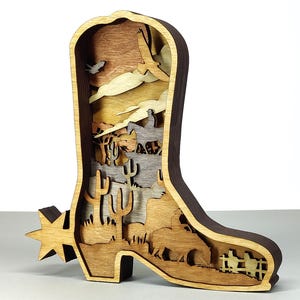 May include: A wooden cowboy boot-shaped art piece with layered desert scene details. The boot features a spur and a layered design depicting cacti, cowboys on horseback, and a soaring bird, all in natural wood tones.