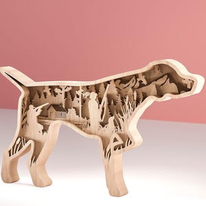 Può includere: Opera d'arte in legno a forma di cane con un design a strati. L'interno raffigura una scena di caccia con alberi, una capanna e figure. I toni naturali del legno contrastano con lo sfondo rosa, creando un oggetto decorativo unico.
