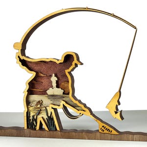 Op de afbeelding: Een gelaagd houten kunstwerk dat een visser toont die een lijn uitwerpt. Het kunstwerk toont een silhouet van een visser, een hengel en een gelaagde scène van een meer met een boot. Het stuk is gemaakt van verschillende bruintinten.