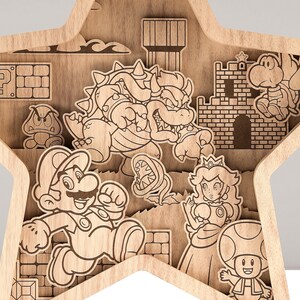 Puede incluir: Arte de pared de madera en forma de estrella con un dise&ntilde;o en capas que presenta personajes de la serie de videojuegos Super Mario. La obra de arte incluye a Mario, la Princesa Peach, Bowser, Goomba y otros elementos ic&oacute;nicos, todo en un acabado de madera natural.
