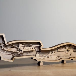 May include: A wooden layered art piece depicting military aircraft and a ship. The artwork is made of light-colored wood and features detailed cutouts of helicopters and a ship, creating a three-dimensional effect. The piece is displayed on a wooden surface.