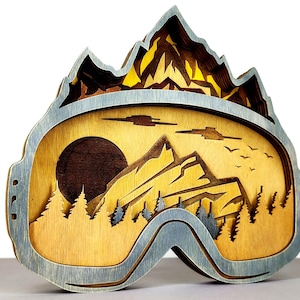 May include: A layered wooden artwork shaped like ski goggles, featuring a mountain landscape with a sunset, trees, and birds. The goggles have a grey frame and a natural wood interior. The mountain peaks are a darker brown.
