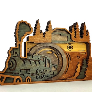 May include: A layered wooden art piece depicting a train and a camera lens with trees. The train is green and brown, with smoke rising from the smokestack. The camera lens is brown and tan, with trees on top.