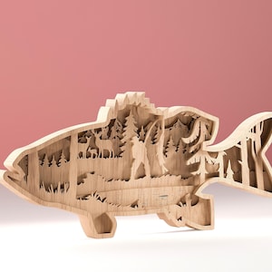 Op de afbeelding: Een houten decoratief stuk in de vorm van een vis met een gelaagd ontwerp. De binnenkant toont een boslandschap met bomen, herten en een wandelaar. Het stuk is gemaakt van lichtgekleurd hout en staat op een wit oppervlak tegen een roze achtergrond.