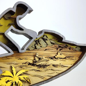 Pu&ograve; includere: Scultura in legno multistrato raffigurante una persona che fa jet ski. La scultura presenta un cielo giallo, acqua marrone e una palma gialla. Il pilota di jet ski &egrave; grigio e la scultura ha un bordo grigio.