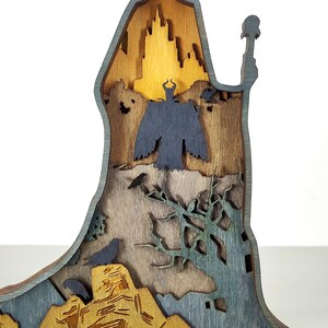 Puede incluir: Una pieza de arte de madera multicapa que presenta una silueta de castillo, una figura oscura y p&aacute;jaros. La pieza est&aacute; hecha de varios tonos de madera y pintada con detalles en azul y verde, creando un efecto dimensional. El tema general es la fantas&iacute;a.
