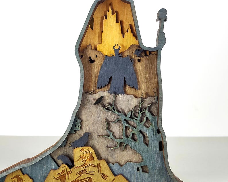 Puede incluir: Una pieza de arte de madera multicapa que presenta una silueta de castillo, una figura oscura y p&aacute;jaros. La pieza est&aacute; hecha de varios tonos de madera y pintada con detalles en azul y verde, creando un efecto dimensional. El tema general es la fantas&iacute;a.