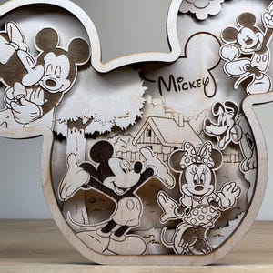 May include: Wooden layered art piece in the shape of Mickey Mouse's head. The artwork features Mickey and Minnie Mouse, Pluto, and a house scene. The word "Mickey" is written on the piece. The art is made of light-colored wood.