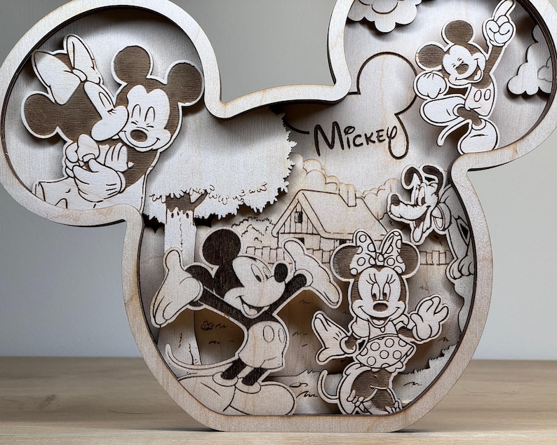 May include: Wooden layered art piece in the shape of Mickey Mouse's head. The artwork features Mickey and Minnie Mouse, Pluto, and a house scene. The word "Mickey" is written on the piece. The art is made of light-colored wood.