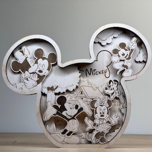 May include: Wooden Mickey Mouse-shaped decorative piece featuring laser-cut images of Mickey, Minnie, Goofy, and other characters. The word "Mickey" is written on the piece. The artwork is in a light brown color.
