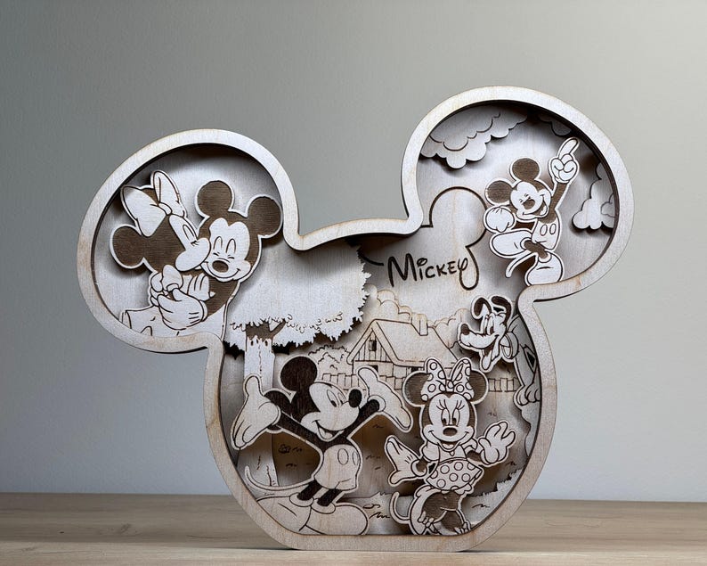 May include: Wooden Mickey Mouse-shaped decorative piece featuring laser-cut images of Mickey, Minnie, Goofy, and other characters. The word "Mickey" is written on the piece. The artwork is in a light brown color.