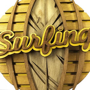 May include: Wooden wall art featuring the word "Surfing" in a layered, cursive font. The design incorporates a surfboard shape with a wood grain finish, set against a circular background with a sunburst effect. The color palette includes shades of brown and yellow.