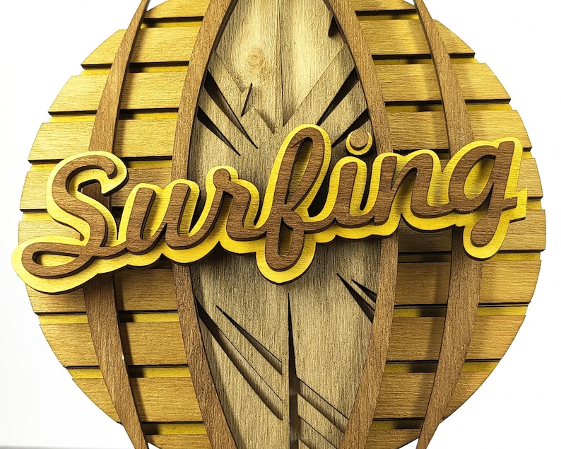 May include: Wooden wall art featuring the word "Surfing" in a layered, cursive font. The design incorporates a surfboard shape with a wood grain finish, set against a circular background with a sunburst effect. The color palette includes shades of brown and yellow.