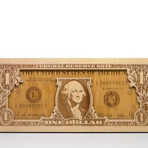 Puede incluir: Una talla de madera de un billete de un dólar. El diseño incluye el texto "THE UNITED STATES OF AMERICA" y "ONE DOLLAR", junto con el retrato de George Washington. La talla es rectangular y de color marrón claro.