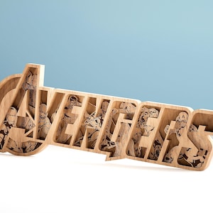 May include: Wooden Avengers sign featuring laser-cut illustrations of various characters. The sign is made of light-colored wood and has a three-dimensional effect, with the characters visible within the letters. The background is a light blue.